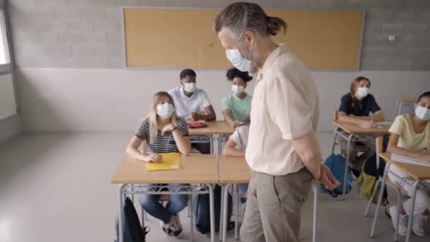 Un enseignant portant un masque de protection debout devant les élèves dans la salle de classe. Expliquer la leçon de son travail à l'école pendant l'épidémie de covidé-19. — Video