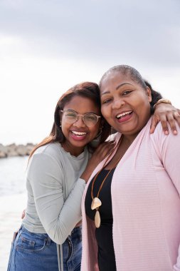 Dikey. Neşeli anne ve kız güneşli bir sahilde kucaklaşıyorlar, neşeli anları paylaşıyorlar ve güneşin sıcaklığının ve doğanın güzelliğinin tadını çıkarırken birlikte anılar yaratıyorlar.