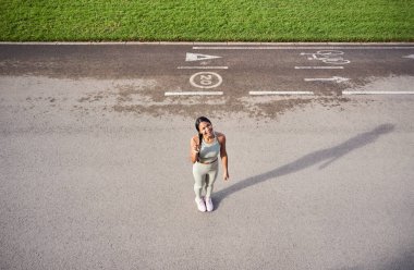 Neşeli Afrikalı bin yıllık kadın spor kıyafetleriyle dışarıda duruyor, gülümsüyor ve barış işareti yapıyor. Üst perspektif pozitifliği, özgürlüğü ve sağlıklı yaşam tarzını vurguluyor.