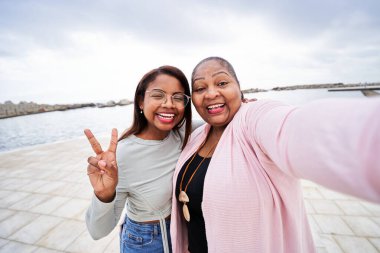 Mutlu anne ve kız deniz kenarında selfie çekiyorlar, kız barış işareti yapıyor, aralarındaki bağı kutluyor ve deniz kenarında tatilin keyfini çıkarıyorlar.