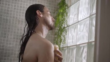 Genç, beyaz bir adam kendini sünger ve jöle duşuyla yıkıyor. Evde banyo işlemlerinin keyfini çıkarıyor. Beyaz milenyumun seçici odak noktası. Uzun saçlı erkek cilt bakımını yapıyor. 