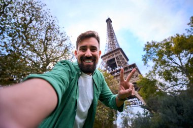 Gülümseyen beyaz adam Paris 'teki Eyfel Kulesi' nde selfie çekiyor ve barış işareti yapıyor. Güneş gözlüğü ve yeşillikle günlük seyahat portresi, doğal ışık altında neşeli şehir atmosferi..