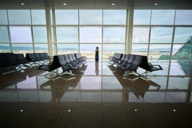 Binlerce yıllık Afrikalı bir kadının havaalanı terminalinde boş koltukları olan büyük bir pencerenin önünde duruşu. Yansımalı simetrik sahne, sakin ortam ve geniş kopya alanı.