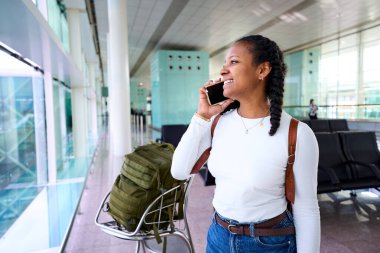 Gülümseyen Afrikalı bin yıllık kadın akıllı telefonuyla bir havaalanı terminalinde sırt çantalı bir tramvayın yanında konuşuyor, seyahat havasını, beklentilerini ve bağlantılarını aktarıyor..