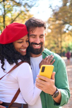 Dikey. Sonbahar parkında sarılıp akıllı telefona bakan mutlu ırklar arası bir çift. Mobil, bağlantı ve neşeli yaşam tarzını kullanarak aşkı aktaran doğal ışık altında yakın plan seyahat portresi.