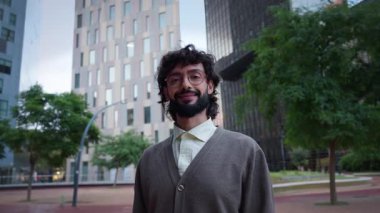 Modern bir iş bölgesinde kamerayla göz teması kuran, gözlüklü, gülümseyen bir milenyum profesyoneli. Arkasındaki şehir merkezi kuleleri güven, liderlik ve kariyer hırsı öneriyor