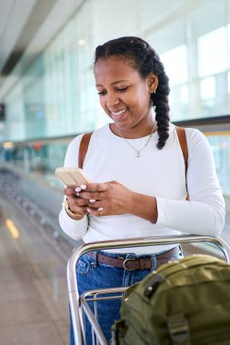 Dikey. Parlak bir havaalanı terminalinde bagaj vagonunu iterken akıllı telefonunu kontrol eden Afrikalı bir kadın. Cam yürüyüş yolu ve tabela biniş zamanı, yolculuk havası ve beklenti öneriyor..