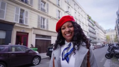 African millennial woman walks along a Paris street wearing a red beret and coat, smiling with direct gaze amid Haussmann buildings, urban travel mood on a cloudy day.