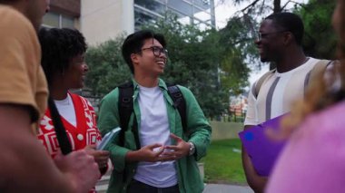 Çeşitli çok ırklı üniversite öğrencileri dersten sonra dışarıda toplanıyor, gülümsüyor ve not defterleri ve sırt çantalarıyla konuşuyorlar. Rahat yeşil bir alanda rahat bir kampüs havası, takım çalışması ve arkadaşlık..
