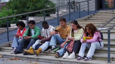 Çeşitli öğrenciler dersten sonra kampüs basamaklarında oturup sohbet ederken ve notları kontrol ederken akıllı telefonlara dalıyorlar. Teknolojik kullanım, eğitim materyalleri ve gevşek bir mola ile gündelik üniversite havası.