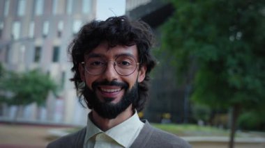 Gözlüklü, gülümseyen bir profesyonel modern kentsel alanda kameraya bakıyor. Güçlü bir göz teması ile yakın çekim güven, yakınlık ve kurumsal bir titreşim taşır..