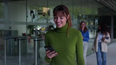 Kafkasya 'nın bin yıllık çalışanı, arka planda iş arkadaşları ve cam parçalarıyla akıllı telefonuna gülümserek modern bir ofis koridorunda yürüyor.