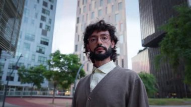 Modern bir iş bölgesinde gözlüklü, kendine güvenen bin yıllık bir adam ciddi ve odaklanmış bir ifadeyle kameraya bakıyor. Arka planda kentsel ofis binaları.