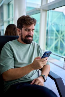 Dikey. Beyaz adam, büyük cam pencerelerin yanındaki bir havaalanı salonunda otururken akıllı telefonunu kontrol ediyor. Rahat seyahat atmosferi, modern mimari ve yatmadan önce yumuşak gün ışığı..