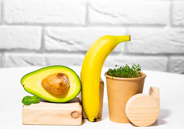 Kreativt stilleben med avokado, banan och mikrogrön på vit bakgrund med träställningar och podium. Jämvikt hälsosam mat. Balanserat näringsbegrepp. — Stockfoto