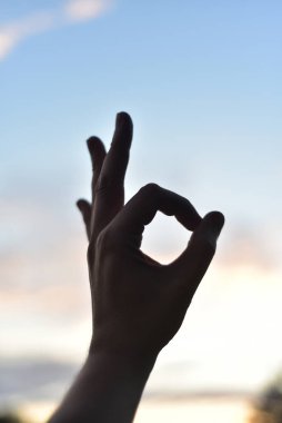 Beyaz bulutlu mavi gökyüzünün arka planında, işaret ve başparmağın halka halinde kapalı olduğunu, diğerlerinin ise yükseldiğini gösterir.