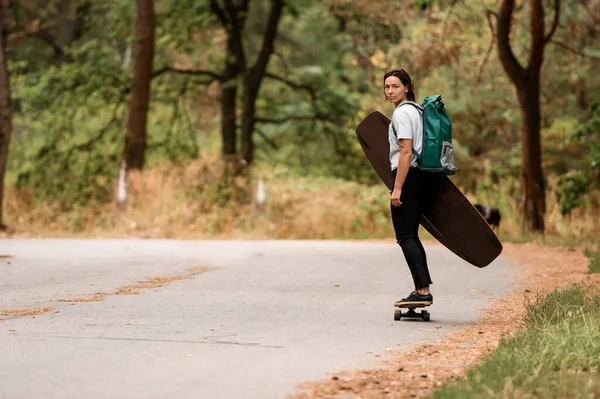 Jonge sportieve vrouw paardrijden skateboard met wakeboard in haar hand en rugzak op haar schouders. — Stockfoto