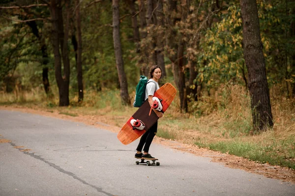 Boční pohled na mladou ženu na koni skateboard s wakeboard v ruce a batoh na ramenou. — Stock fotografie