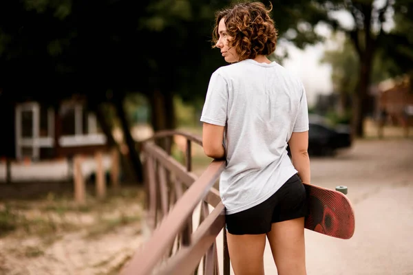 Mooie jonge vrouw met krullend haar in wit t-shirt houdt skateboard — Stockfoto