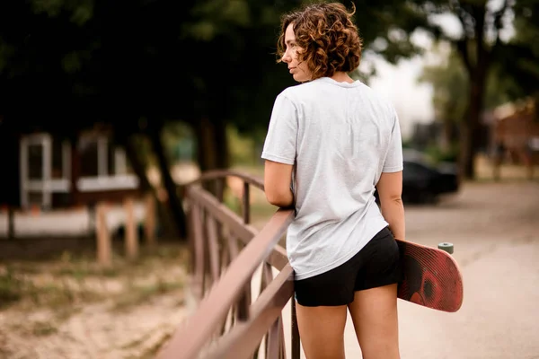 Bela jovem mulher com cabelo encaracolado em t-shirt branca detém skate — Fotografia de Stock