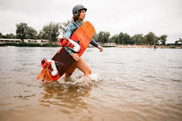 Schöne Frau mit Helm und Wakeboarderbrett spaziert auf dem Wasser am Ufer des Flusses entlang — Stockfoto