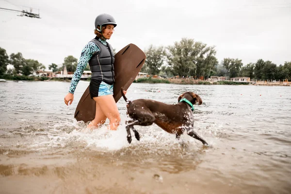 若いです女性でヘルメットとウェットスーツ歩くとともに陽気な犬に沿って川の水 — ストック写真