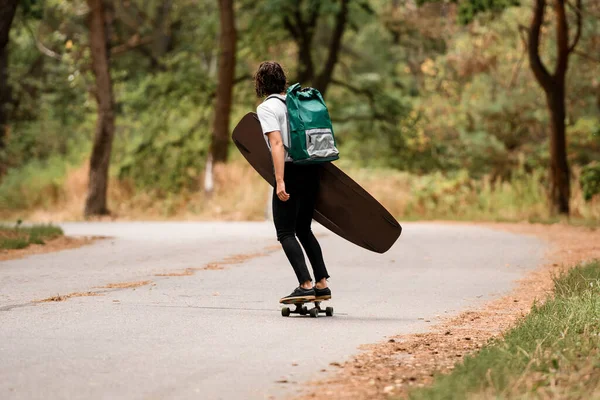 Πίσω όψη της νεαρής γυναίκας ιππασία skateboard με wakeboard στο χέρι και σακίδιο στους ώμους της. — Φωτογραφία Αρχείου