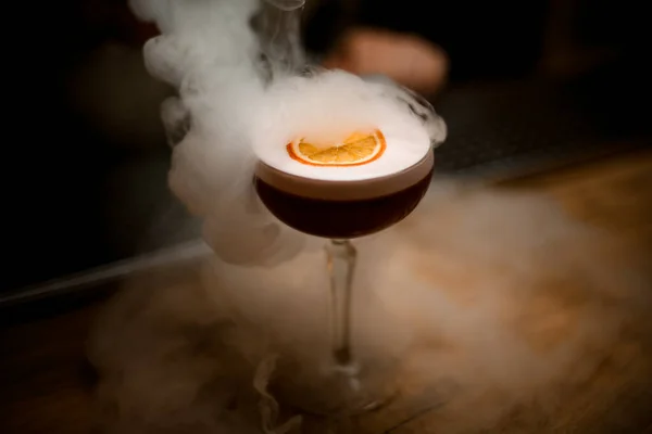Primo piano di vetro di bevanda con schiuma e fetta di stand di agrumi su bancone di bar fumoso. — Foto Stock