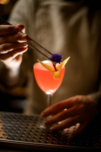Homme barman décore soigneusement verre de vin de cocktail lumineux avec de la fleur à l'aide de pinces — Photo