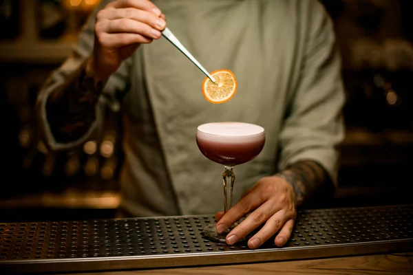 Bartender holds tweezers with slice of orange over wine glass with foam cocktail — Stock Photo, Image