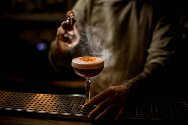 Mannelijke barman nauwkeurige hagelslag op cocktailglas met schuim versierd met sinaasappel en kaneel — Stockfoto