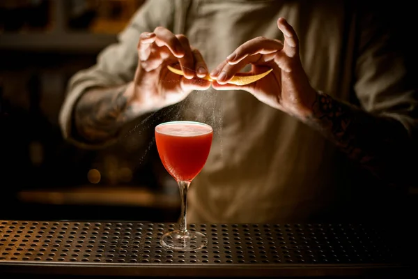 Man barman houdt citroenschil en hagelslag op glas met heldere cocktail. — Stockfoto