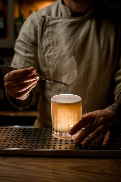 Man bartender håller pincett med dekorativa blad för att dekorera glas med cocktail — Stockfoto