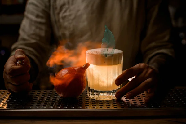 Barman opatrně stříkne a zapálí malou dýni. Sklenice nápoje stojí poblíž — Stock fotografie