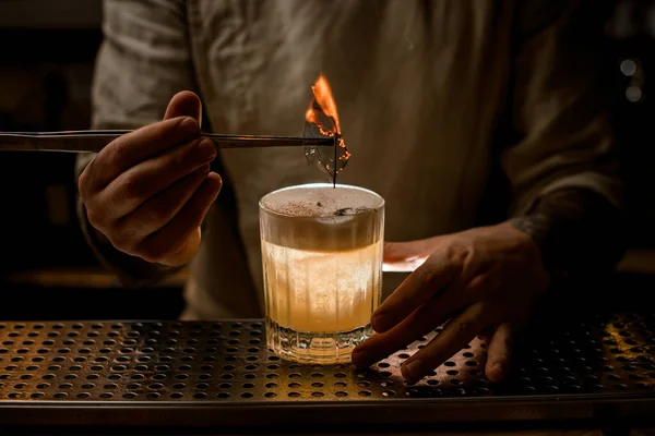 Бармен держит пинцет с горящим листом, чтобы украсить стакан коктейля — стоковое фото