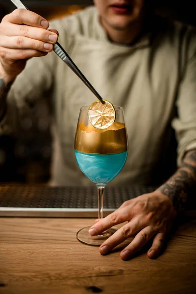 Bartender holds slice of citrus over wine glass decorated with gold — Stock Photo, Image