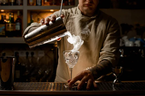 Barkeeper gießt dampfenden Drink aus Stahlbecher in Weinglas mit Eiswürfeln. — Stockfoto