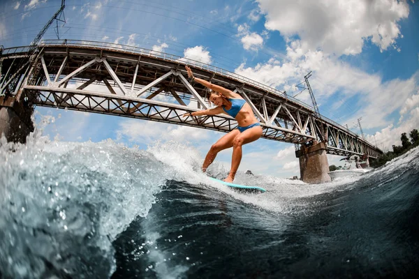 도시의 다리 배경에 높은 파도가 치는 판위에서 균형을 잡고 있는 운동 선수 여자. — 스톡 사진