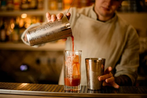 Homem bartender detém vidro agitador de aço e derrama coquetel vermelho a partir dele em outro vidro. — Fotografia de Stock