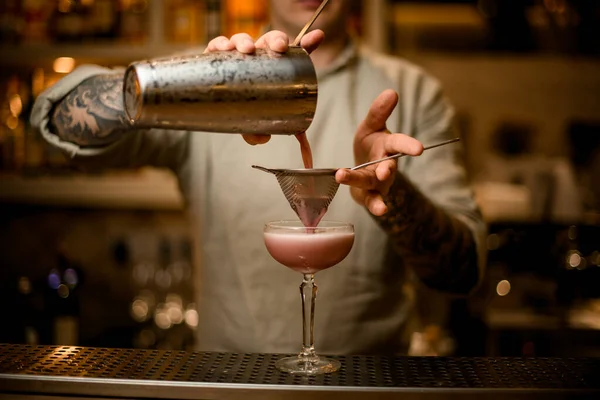 Bartender holds sieve over wine glass and pours pink cocktail from shaker. — Stock Photo, Image