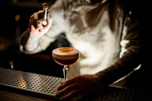Barman strooit voorzichtig op cocktailglas met schuim versierd met sinaasappel en kaneel — Stockfoto