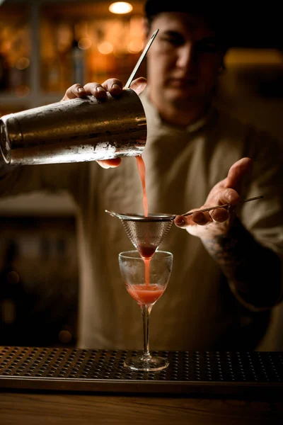 Νεαρός άνδρας μπάρμαν κρατά κόσκινο πάνω από ποτήρι κρασιού και απαλά ρίχνει κοκτέιλ από αναδευτήρα. — Φωτογραφία Αρχείου