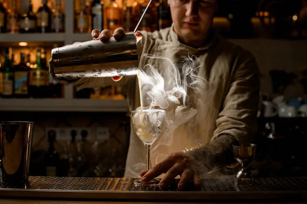 Mannelijke barman schenkt stomende drank uit stalen shaker cup in wijnglas met ijsblokjes. — Stockfoto