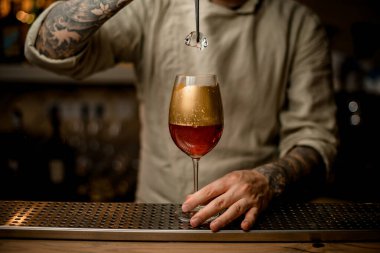 Barmen bir parça buzu bardağın üzerinde tutuyor. Altınla süslenmiş köpüklü bir içecek.