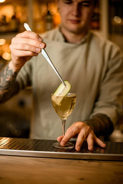 Hombre en el bar decora copa de cóctel con rodaja de pepino con pinzas —  Fotos de Stock