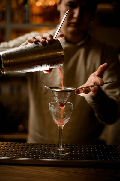 Бармен держит решето над бокалом вина и нежно наливает коктейль из шейкера. — стоковое фото