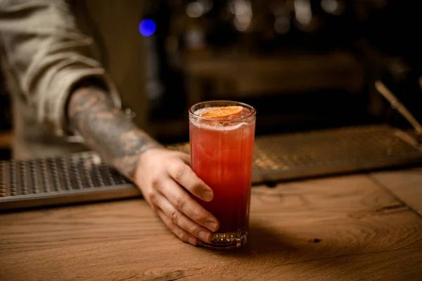 Barmans hand houdt glas met koude drank met ijsblokjes, versieren met sinaasappelschijfje — Stockfoto