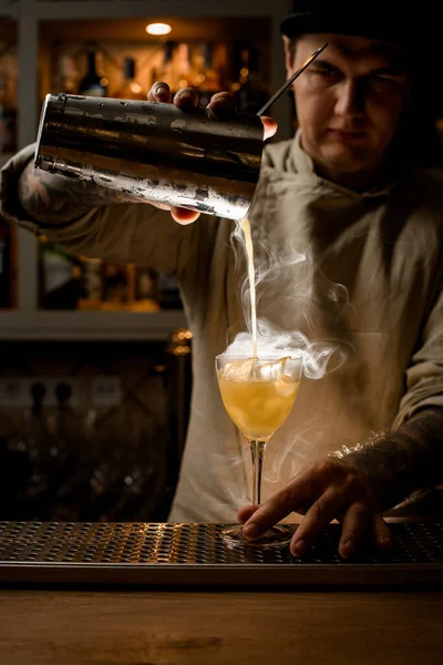 Człowiek nalewa parujący napój ze stalowego shakera do kieliszka do wina z kostkami lodu. — Zdjęcie stockowe