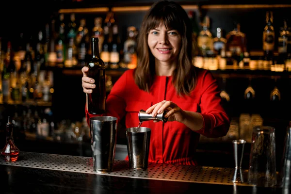 Smiling woman bartender pours liquid from jigger into steel shaker cup standing on bar — Stock Photo, Image