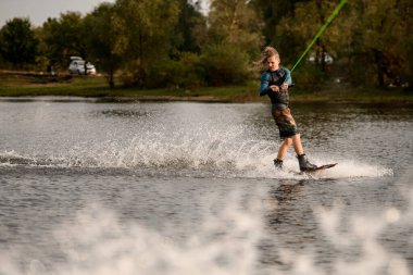 Dalgıç kıyafetli ve yelekli genç adam nehirde wakeboard sürüyor.